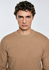 Giovane uomo con capelli ricci, che indossa un maglione beige a trama, guarda direttamente nella macchina fotografica su uno sfondo neutro.