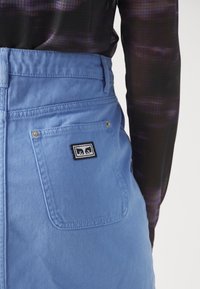Gros plan sur une personne portant un pantalon en denim bleu clair avec un patch logo carré sur la poche arrière et un haut à manches longues transparent foncé.