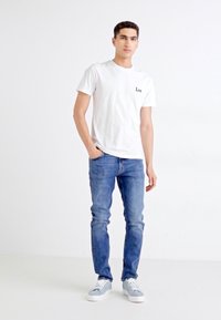 Camiseta blanca de algodón con logo "Lee", jeans de mezclilla azul y zapatillas gris claro. El modelo está de pie frente a un fondo liso.