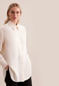 Blusa a maniche lunghe color crema chiaro con chiusura a bottoni, colletto, spacchi laterali e tessuto liscio, abbinata a pantaloni neri.