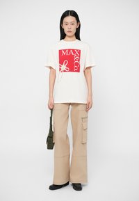 Γυναίκα με λευκή oversized μπλούζα με κόκκινη εκτύπωση "MAX&Co.", μπεζ φαρδιά cargo παντελόνια, μαύρα μποτίνια και κρατώντας μια μικρή πράσινη τσάντα.