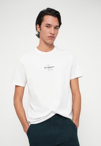 Mladý muž s tmavými vlasmi, ktorý má na sebe biele tričko Calvin Klein a tmavé nohavice, stojí so založenými rukami v vreckách na jednoduchom pozadí.