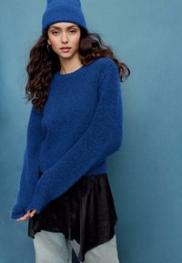 Modrý chlpatý sveter s dlhými rukávmi a rebrovaným lemom, navrstvený na čiernom saténovom topánke s plynúcim dizajnom. Štylizovaný s svetlomodrými džínami.