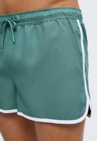 Πράσινο μαγιό σορτς με λευκή επένδυση και κορδόνι στη μέση, φορεμένο στο κάτω μέρος της κοιλιάς και στο πάνω μέρος του μηρού ενός ατόμου.