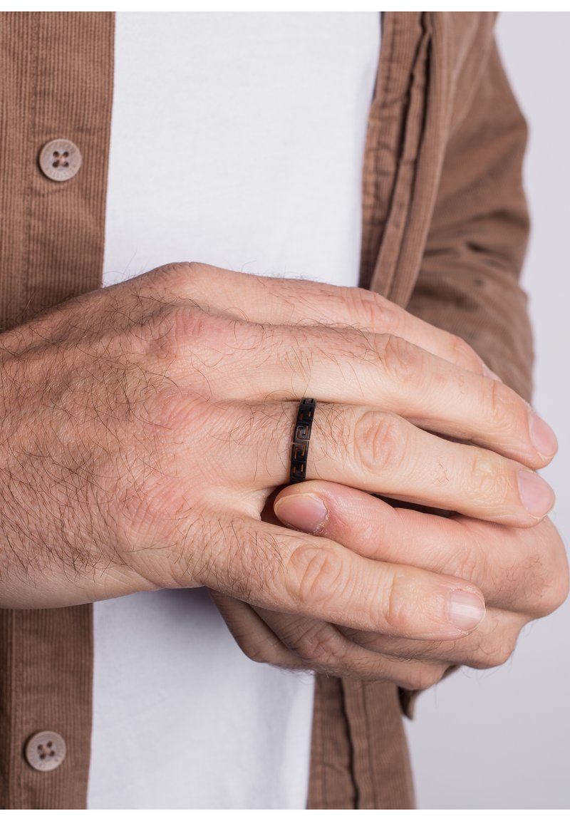 Black metal ring with a patterned design, worn on a hand with light skin. The hand is positioned against a brown fabric background.
