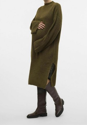 Een zwangere persoon die een oversized olijfgroene gebreide jurk met wijde mouwen draagt en bruine kniehoge laarzen, poserend in zijaanzicht.