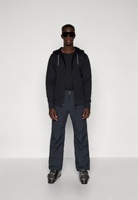 Zwarte hoodie met rits over een zwart shirt, gecombineerd met donkere broek. Draagt een zonnebril en zwarte skischoenen met metalen gespen.