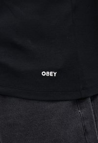 Μαύρο ριμπ μπλουζάκι με τη λέξη "OBEY" σε λευκή κεντητική στη βάση, συνδυασμένο με σκούρο γκρι υφασμένo παντελόνι.