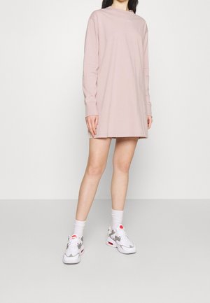 Vrouw draagt een lichtroze oversized jurk met lange mouwen, witte en grijze sneakers en witte sokken, staand tegen een effen witte achtergrond.