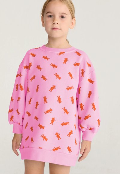 Jeune fille portant un sweat-shirt rose oversize avec des motifs d’ours orange, debout devant un fond clair uni.