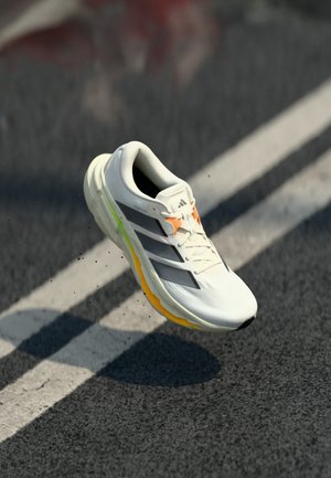 Weißer und schwarzer Adidas-Laufschuh mit orangefarbenen und gelben Details, schwebt über einer Asphaltstraße mit weißen Fahrbahnmarkierungen.
