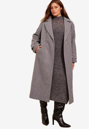 Manteau en mélange de laine gris avec des revers surdimensionnés, double boutons et poches avant, associé à une robe gris foncé et des bottes à talons noirs brillantes.