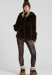 Brun fuskpälsjacka med dragsko i midjan, kombinerad med leggings i svart och brunt leopardmönster. Modellen bär en brun stickad mössa.