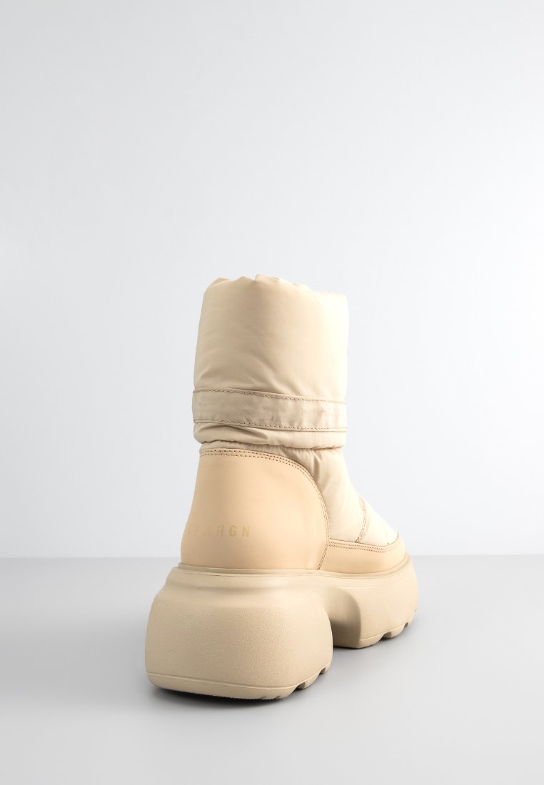 Beige gepolsterter Knöchelstiefel mit einer dicken Gummisohle und glatter Textur. Verfügt über eine abgerundete Form und dezente Markenkennzeichnung an der Seite.