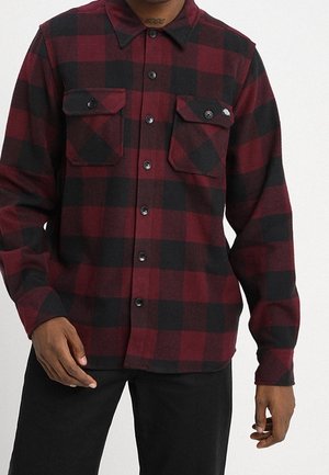 Mann trägt ein rot-schwarz kariertes Hemd mit Knöpfen und zwei Brusttaschen, kombiniert mit einer schwarzen Hose.