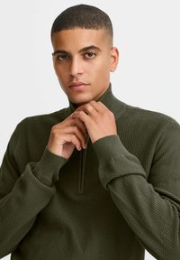 Olivgrüner Pullover mit einem halben Reißverschluss am Kragen. Hergestellt aus einem strukturierten Stoff mit einem gestrickten Muster. Enganliegende Bündchen und Körper.