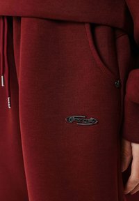 Bordeauxrode joggingbroek met een zachte textuur, voorzien van een zijzak en een zilveren logopatch. Trekkoorddetail in de taille.