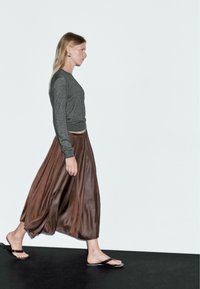Vrouw die loopt en een donkergrijs langemouwen top, een bruine vloeiende midi-jurk, een paar grote oorbellen en zwarte slippers draagt, tegen een eenvoudige witte achtergrond.