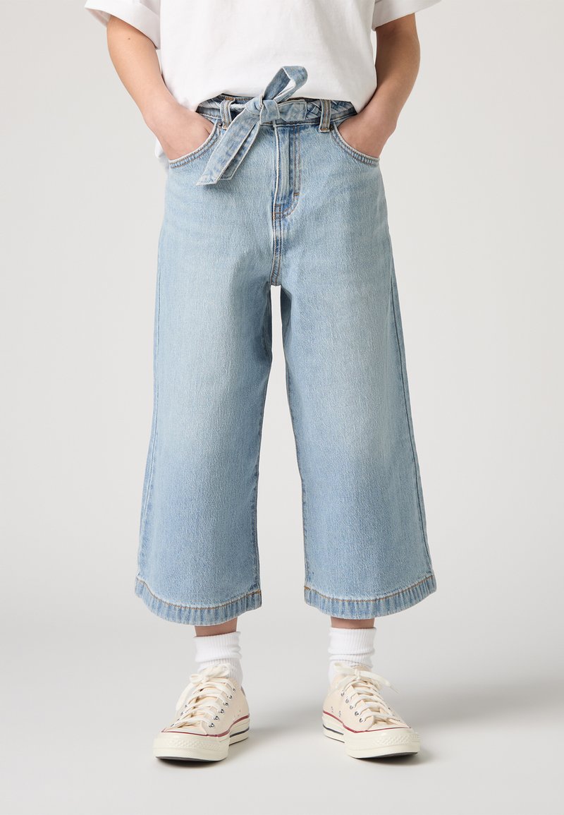 Pantaloni in denim azzurro chiaro, a gamba larga e con vita annodata, taglio cropped e orlo sfrangiato, abbinati a sneakers bianche e calze.