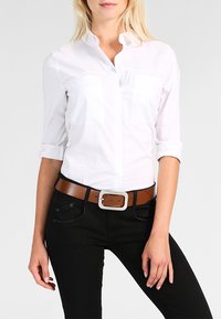 Chemise blanche à manches longues avec deux poches poitrine, associée à un pantalon noir et une ceinture en cuir marron avec des accessoires en argent.