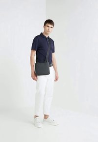 Borsa a tracolla nera in materiale testurizzato, forma quadrata, con tracolla regolabile. Indossata con polo blu navy e pantaloni bianchi, in piedi su sfondo chiaro.