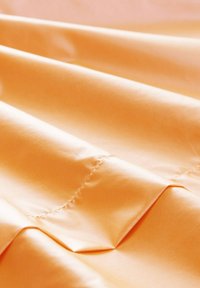 Tessuto di raso in una delicata tonalità arancione, con una consistenza liscia e un leggero lucido, bordi piegati eleganti e dettagli di cucitura diritta.