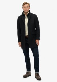 Abrigo de lana negro con cuello con muescas, botones frontales y bolsillos laterales sobre un suéter color crema, combinado con jeans oscuros y botas marrones.