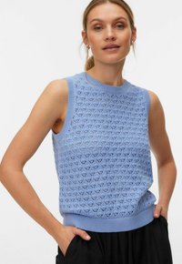 Femme portant un haut en tricot bleu clair sans manches avec un motif en diamant, debout les mains dans les poches devant un fond uni.