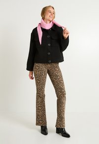 Veste noire avec poches avant, associée à un pantalon évasé en imprimé léopard marron. Écharpe rose drapée autour du cou. Bottes à talons noires.