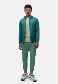 Lettvekts teal jakke med hette, sammen med en lys grønn t-skjorte og matchende smale bukser. Sporty sko med grå og blå detaljer.
