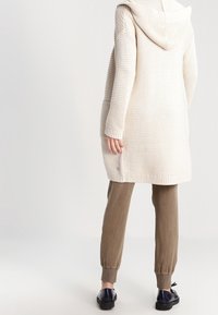 Cárdigan largo de punto color crema con capucha, que presenta puños de canalé y una tela texturizada. Combinado con pantalones ajustados de color marrón claro.