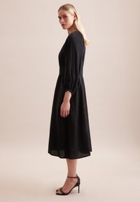 Abito midi nero a maniche lunghe con maniche a sbuffo e vita aderente. Mostra anche una modella che indossa sandali neri con tacco alto.