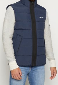 Marineblauer Steppweste mit gestepptem Design, schwarzem Reißverschluss und Seitentaschen. Verfügt über graue Sweatshirtärmel und gerippte Bündchen.