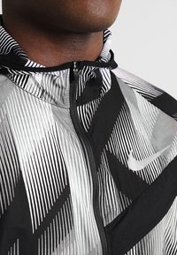 Nike-jakke med et sort og hvidt geometrisk mønster, med lynlåsåbning foran og hætte. Letvægtsstof med en glat tekstur.