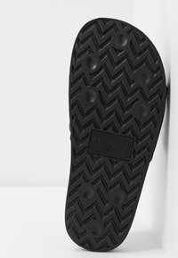 Semelle de chaussure en caoutchouc noir avec un motif de bande de roulement en zigzag, des grips circulaires et un logo Levi's rectangulaire au centre.