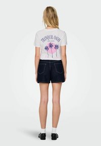 Balts T-krekls ar "Tranquil Oasis" grafiku, kurā ir rozā un zili palmu attēli. Apvienots ar tumšām džinsu šortiem un melnām kurpēm.