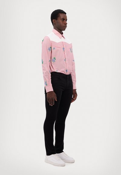 Kariertes Hemd in Pink und Weiß mit floralem Stickmuster und langen Ärmeln, kombiniert mit schwarzen Slim-Fit-Jeans und weißen Sneakers.