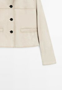 Beige mockasinjacka med krage, fyra svarta knappar, sidfickor och en kort silhuett. Slät textur och jämn sömnad.