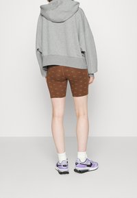 Pantalones cortos de bicicleta marrones con un sutil patrón de logo, combinados con una sudadera gris de gran tamaño y zapatillas moradas con acentos negros.