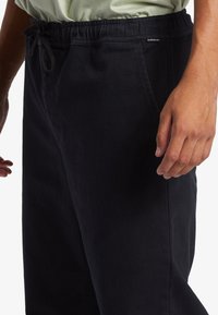 Prim-plan al unei persoane îmbrăcate cu pantaloni negri cu șnur la brâu și buzunare, stând cu o mână relaxată lângă corp.