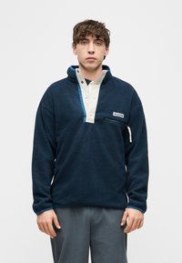 Νέος άνδρας που φοράει μπλε σκούρο φλις πουλόβερ με μπεζ πατιλέτα με κουμπιά και γκρι παντελόνι, στέκεται μπροστά σε απλό ανοιχτό φόντο.