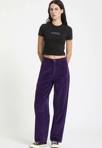 T-shirt noir raccourci avec texte violet, associé à un pantalon large en velours côtelé violet taille haute. La tenue est complétée par des baskets noires.