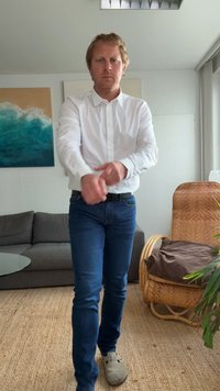Pessoa vestindo uma camisa branca de botões e jeans escuros, com as mãos posicionadas em direção aos pulsos. O ambiente inclui um sofá cinza e plantas de interior.
