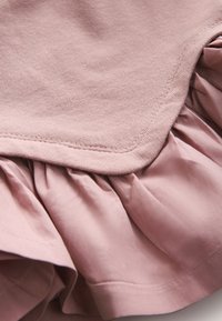 Lähikuva pehmeästä vaaleanpunaisesta kankaasta, jossa näkyy kerroksittain laskostettu kangas, ompeleet ja sileä, mattapintainen rakenne.