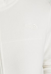 Nahaufnahme einer weißen Fleecejacke mit geprägt The North Face-Logo auf der Brust und einem vertikalen Reißverschlussdetail, das auf der linken Seite sichtbar ist.