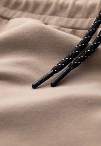 Beige bomullstyg med mjuk textur, prytt med svarta dragsnören, accentuerat med vit sömnad och plastspetsar.