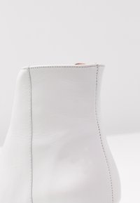 Bottine en cuir blanc avec une texture lisse, une silhouette incurvée et des coutures contrastées le long des coutures. Design minimaliste.