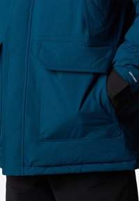 Teal vattentät jacka med en framficka, mjuk textur, justerbara ärmslut och "DRYVENT"-logotyp på ärmen. Har en rak nederkant.