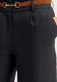 Schwarze Leinenhosen mit einer strukturierten Oberfläche, ausgestattet mit Falten und Seitentaschen, ergänzt durch einen braunen Wildledergürtel mit goldenen Beschlägen.