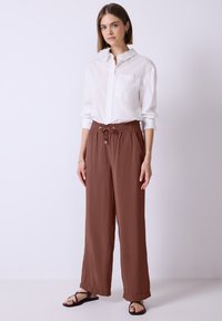 Losse bruine broek met een gestructureerde afwerking en een elastische tailleband, gecombineerd met een wit overhemd met knopen en zwarte sandalen.